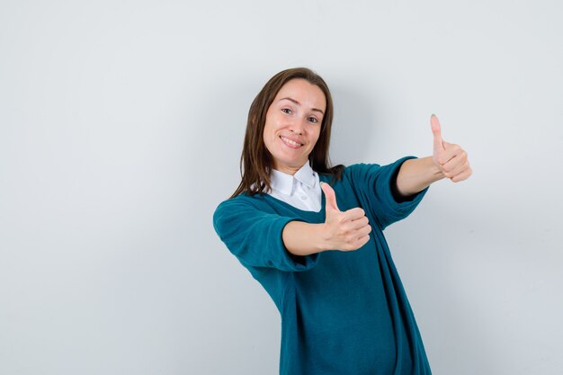 Portret van een jonge vrouw die dubbele duimen in een trui over een wit overhemd laat zien en er vrolijk vooraanzicht uitziet