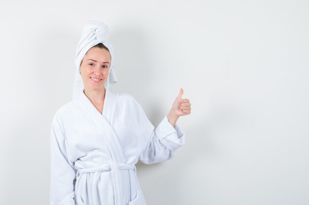 Portret van een jonge vrouw die duim in witte badjas, handdoek toont en vrolijk vooraanzicht kijkt