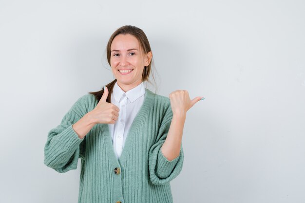 Portret van een jonge vrouw die duim omhoog laat zien, opzij wijst in blouse, vest en er vrolijk vooraanzicht uitziet