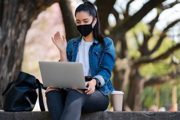 Portret van een jonge vrouw die een gezichtsmasker draagt tijdens een videogesprek met een laptop terwijl ze buiten zit. Stedelijk begrip. Nieuw normaal levensstijlconcept.