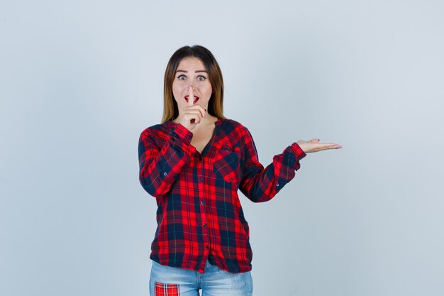 Portret van een jonge vrouw die een stiltegebaar toont, de handpalm opzij spreidt in een geruit overhemd en er opgewonden uitziet