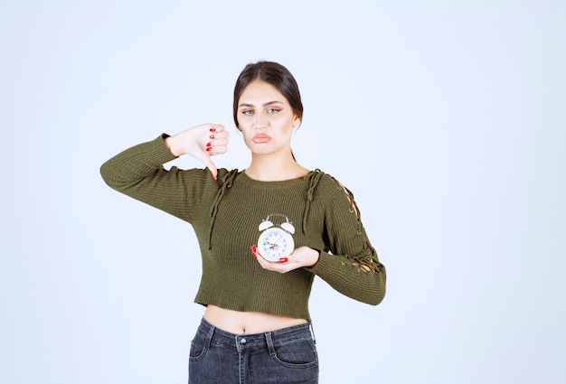 Portret van een jonge vrouw die een wekker vasthoudt en duimen naar beneden geeft.