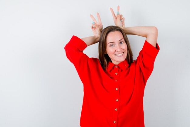 Portret van een jonge vrouw die het gebaar van konijnenoren in een rode blouse toont en er gelukkig vooraanzicht uitziet