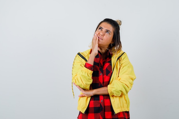 Portret van een jonge vrouw die in een denkende pose staat terwijl ze opkijkt in een geruit hemd, een jas en een verbaasd vooraanzicht kijkt