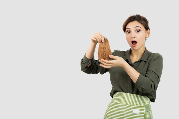 Portret van een jonge vrouw die in schort een kokosnoot houdt tegen witte muur