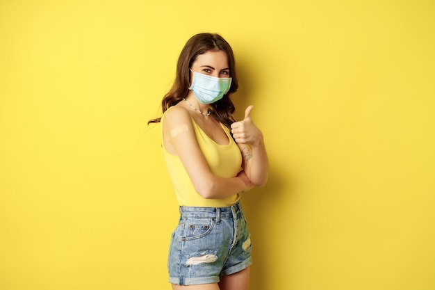Portret van een jonge vrouw die is ingeënt tegen covid-griep met een gezichtsmasker met duimen omhoog, meisje heeft p...