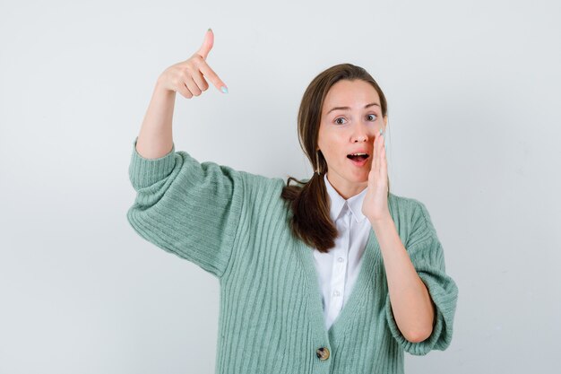 Portret van een jonge vrouw die naar beneden wijst, de hand in de buurt van de mond houdt in een blouse, een vest en een verbaasd vooraanzicht kijkt