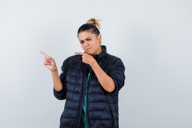 Portret van een jonge vrouw die naar de linkerbovenhoek in een pufferjack wijst en besluiteloos vooraanzicht kijkt