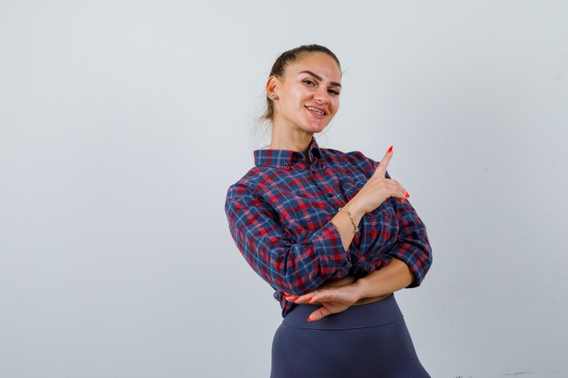 Portret van een jonge vrouw die naar de rechterbovenhoek wijst in een geruit hemd, een broek en een gelukkig vooraanzicht kijkt