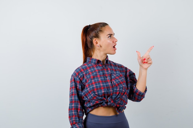 Portret van een jonge vrouw die naar de rechterbovenhoek wijst in een geruit hemd, een broek en een verbaasd vooraanzicht kijkt