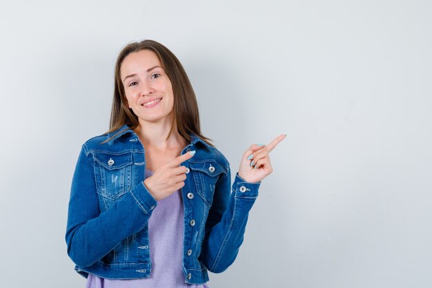 Portret van een jonge vrouw die naar de rechterbovenhoek wijst in t-shirt, jas en er vrolijk vooraanzicht uitziet