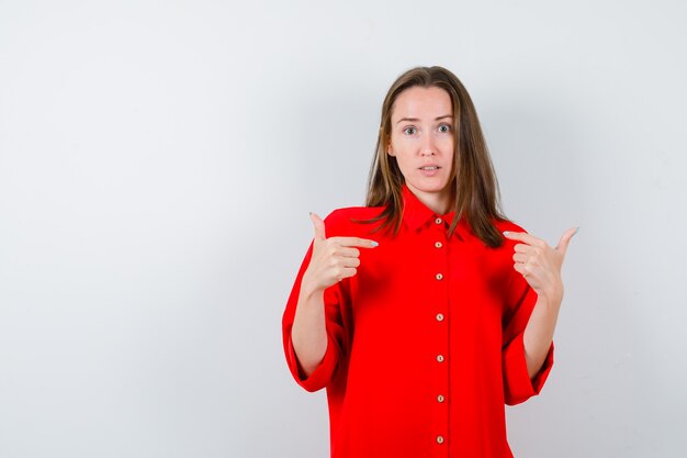 Portret van een jonge vrouw die naar zichzelf wijst in een rode blouse en een verbaasd vooraanzicht kijkt