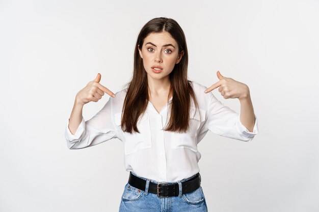 Portret van een jonge vrouw die naar zichzelf wijst, zichzelf bevordert, op een witte achtergrond staat. Ruimte kopiëren