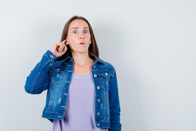 Portret van een jonge vrouw die oor met vinger in t-shirt, jas stopt en zich afvraagt vooraanzicht