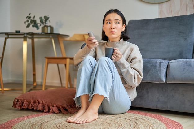 Portret van een jonge vrouw die op afstand tv kijkt en van kanaal wisselt op televisie zittend op de vloer nea