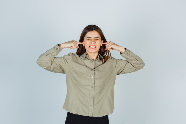 Portret van een jonge vrouw die oren inplugt met vingers in shirt, rok en er geïrriteerd vooraanzicht uitziet