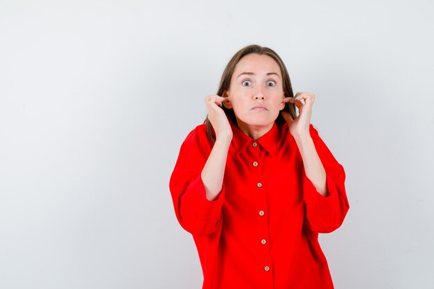 Portret van een jonge vrouw die oren stopt met vingers in een rode blouse en er bang vooraanzicht uitziet