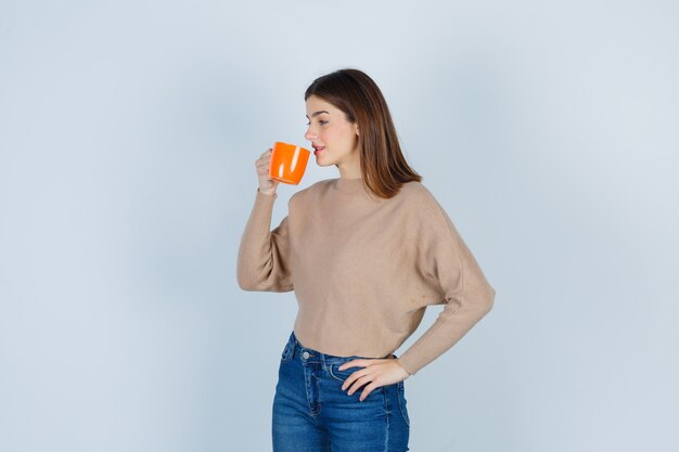 Portret van een jonge vrouw die uit de beker drinkt, met de hand op de taille, zijwaarts in een trui, spijkerbroek en er tevreden uit.