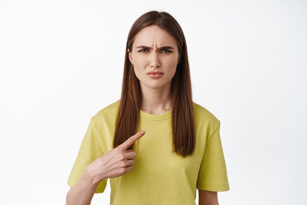 Portret van een jonge vrouw die verward kijkt terwijl ze naar zichzelf wijst, met een casual geel t-shirt, witte achtergrond