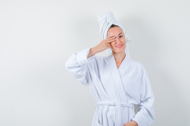 Portret van een jonge vrouw die vingers op oog in witte badjas, handdoek houdt en vrolijk vooraanzicht kijkt