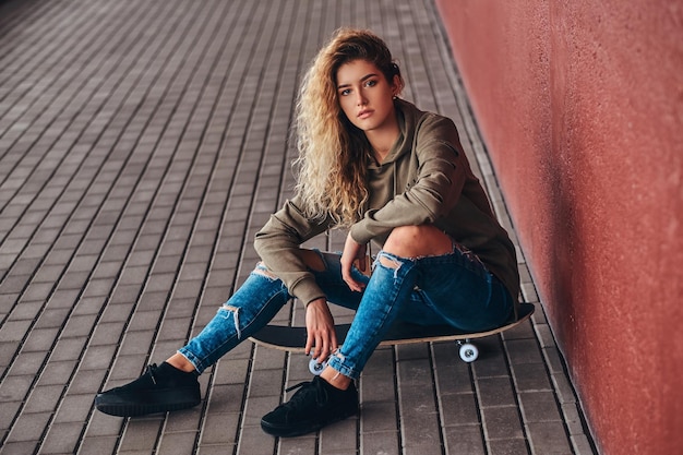 Portret van een jonge vrouw gekleed in een hoodie en gescheurde spijkerbroek zittend op een skateboard bij een brugvoetgang.