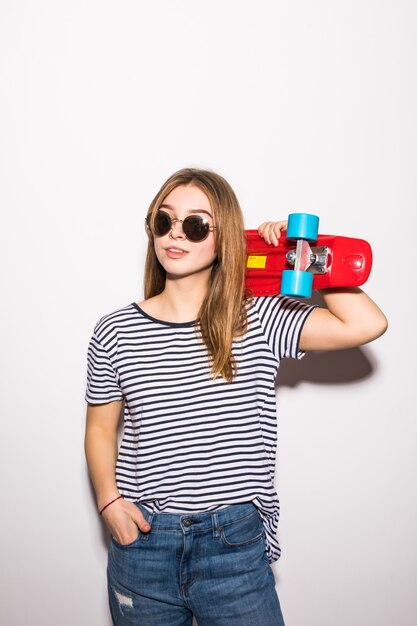 Portret van een jonge vrouw in zonnebril die met skateboard stellen terwijl status over witte muur