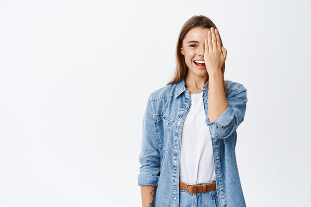 Portret van een jonge vrouw met blond haar, bedek de helft van het gezicht met make-up met natuurlijk licht, laat zien voor het effect of controleer het zicht in de opticienwinkel, witte muur