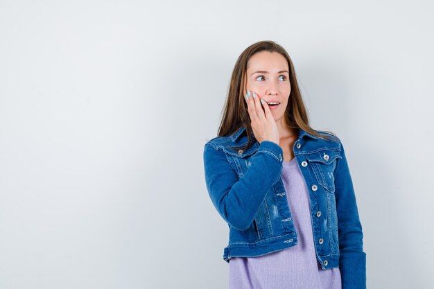 Portret van een jonge vrouw met de hand op de wang, wegkijkend in t-shirt, jas en verbaasd vooraanzicht