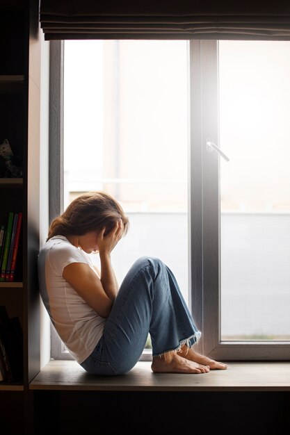 Portret van een jonge vrouw met een laag zelfbeeld die thuis bij het raam zit