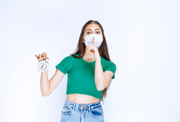 Portret van een jonge vrouw met een medisch masker die een wekker houdt.