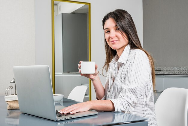 Portret van een jonge vrouw met koffiekopje zit aan ontbijttafel met laptop