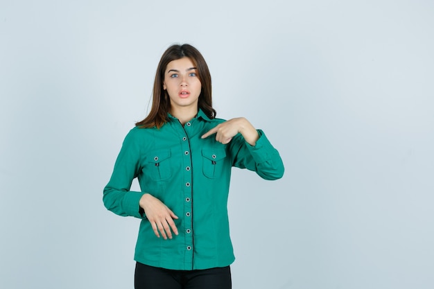 Portret van een jonge vrouw wijzend op zichzelf in een groen shirt en verbaasd vooraanzicht op zoek