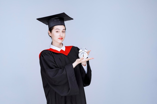 Portret van een jonge vrouwelijke afgestudeerde student die de klok vasthoudt om de tijd te tonen. Hoge kwaliteit foto