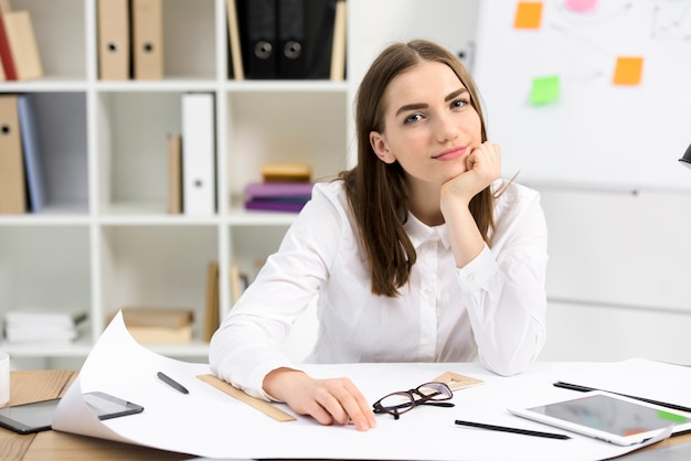 Portret van een jonge vrouwelijke architectenzitting bij bureau die aan camera kijken