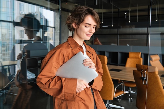 Gratis foto portret van een jonge vrouwelijke programmeur, een digitale nomade die op kantoor werkt en met haar laptop staat