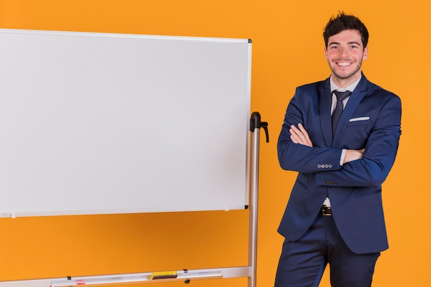 Gratis foto portret van een jonge zakenman met zijn gekruiste arm staande in de buurt van het whiteboard tegen een oranje achtergrond