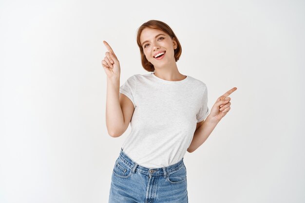 Portret van een jonge zorgeloze vrouw die naar twee kanten wijst. Meisje dat links en rechts manieren toont, op een witte muur staat en glimlacht