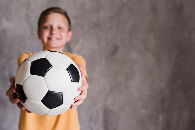 Portret van een jongen die voetbalbal naar camera tonen die zich voor van concrete muur bevinden