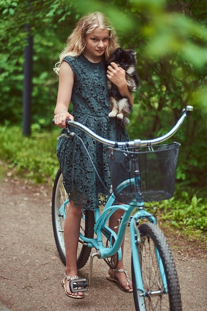 Portret van een klein blond meisje in een casual jurk, houdt een schattige spitzhond vast. rijd op een fiets in een park.