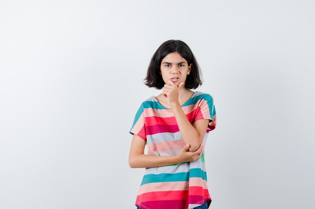 Portret van een klein meisje dat de hand op de kin in een t-shirt houdt en van voren overstuur kijkt