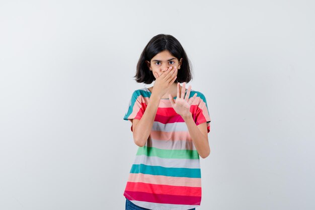 Portret van een klein meisje dat de hand op de mond houdt, een stopgebaar in een t-shirt laat zien en een bang vooraanzicht ziet