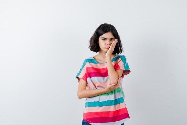 Portret van een klein meisje dat de hand op de wang in een t-shirt houdt en van voren overstuur kijkt