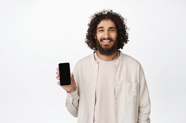 Portret van een knappe Arabische man die het scherm van een mobiele telefoon toont en glimlacht en een witte achtergrond van de toepassing aanbeveelt