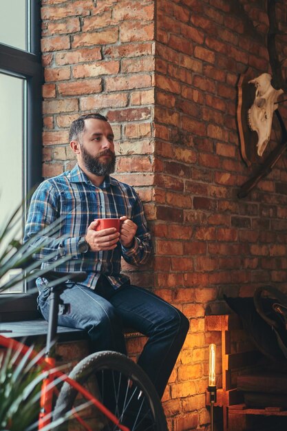 Portret van een knappe bebaarde hipster man in een blauw fleece shirt en spijkerbroek houdt een kopje koffie in de ochtend zittend op een vensterbank in een studio met loft interieur.