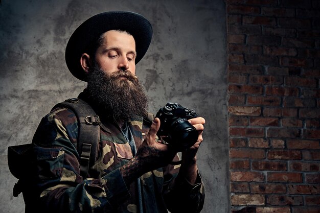 Portret van een knappe bebaarde reiziger in een hoed en camouflagejas, met een rugzak en getatoeëerde armen, houdt een fotocamera vast. Geïsoleerd op een donkere achtergrond.