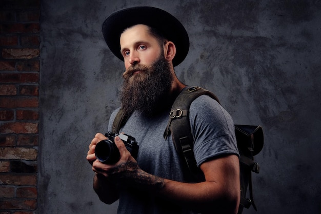 Portret van een knappe bebaarde reiziger in een hoed met een rugzak en getatoeëerde armen, houdt een fotocamera vast. Geïsoleerd op een donkere achtergrond.