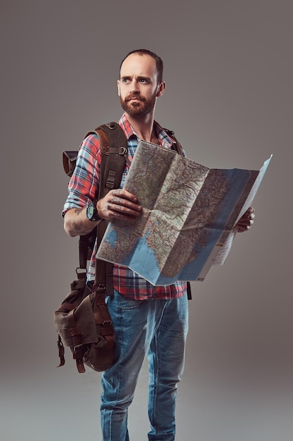 Portret van een knappe getatoeëerde reiziger in een flanellen shirt met een rugzak, houdt een kaart vast, staande in een studio. Geïsoleerd op een grijze achtergrond.