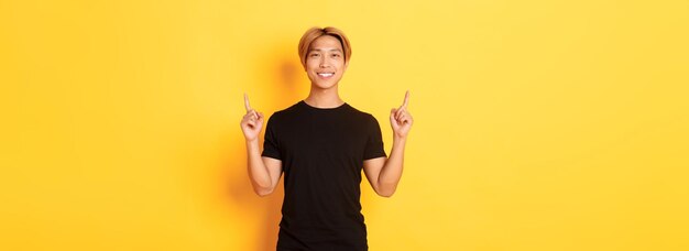 Portret van een knappe glimlachende aziatische man in een zwart t-shirt die met zijn vingers omhoog wijst en er tevreden uitziet