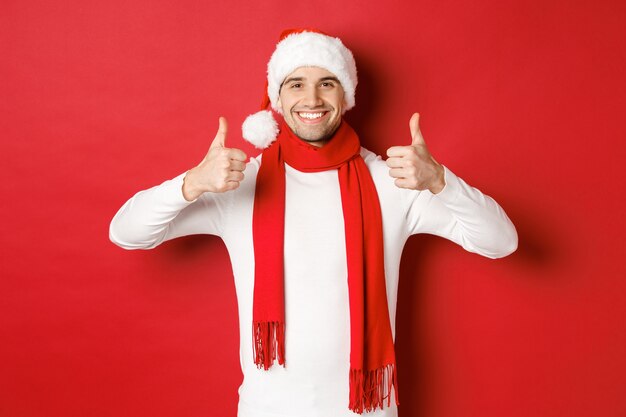 Portret van een knappe Glimlachende man in een kerstsjaal en muts met thumbsup die de kerststand viert...