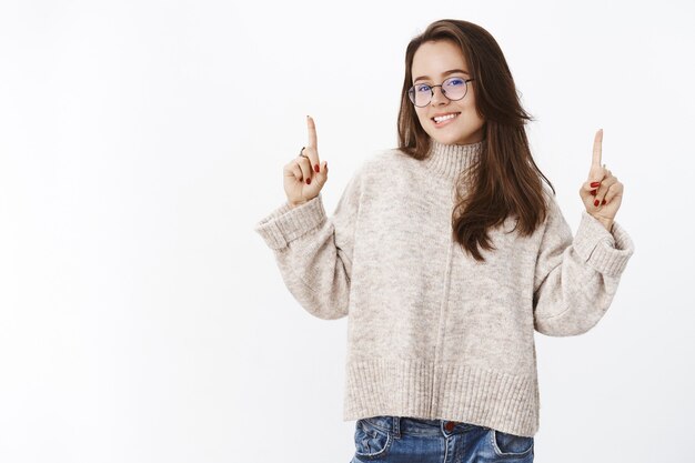 Portret van een knappe jonge en zelfverzekerde vrouw met een bril en een warme trui die handen opsteekt, glimlachend en bijtend op de onderlip van verrukking en verlangen, gretig probeer het product zelf over de grijze muur.